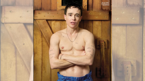 Shirtless man with arms crossed stands against a wooden wall, tattoos visible on his chest and arms, wearing blue striped shorts.