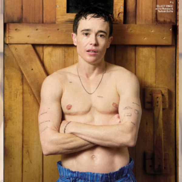 Shirtless man with arms crossed stands against a wooden wall, tattoos visible on his chest and arms, wearing blue striped shorts.