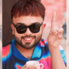 Smiling man in sunglasses makes a peace sign with two fingers, holding a cup in a colorful shirt.