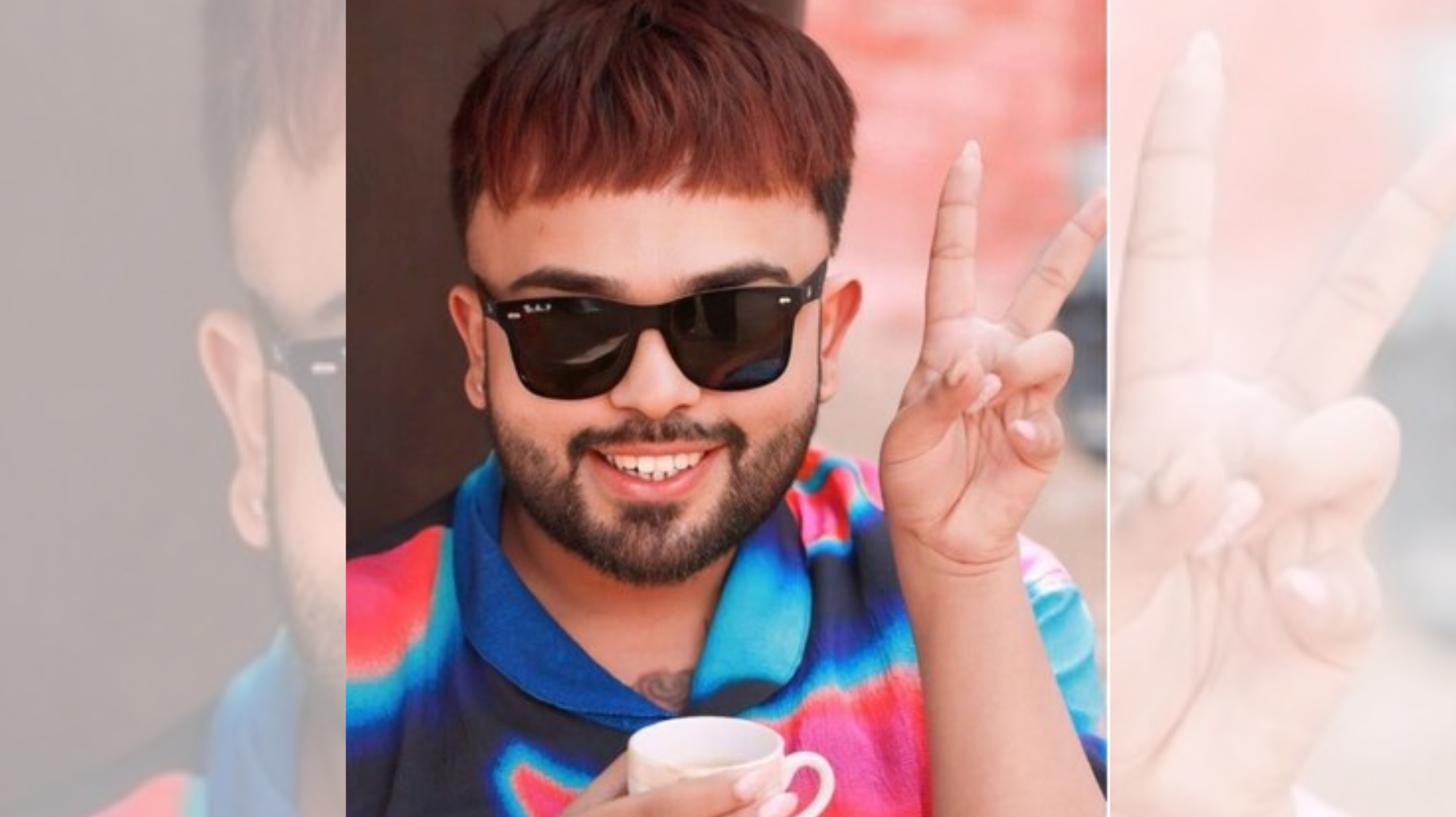 Smiling man in sunglasses makes a peace sign with two fingers, holding a cup in a colorful shirt.