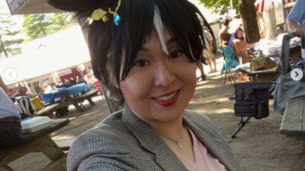 Smiling woman in a gray blazer takes a sunny selfie at an outdoor market or festival.