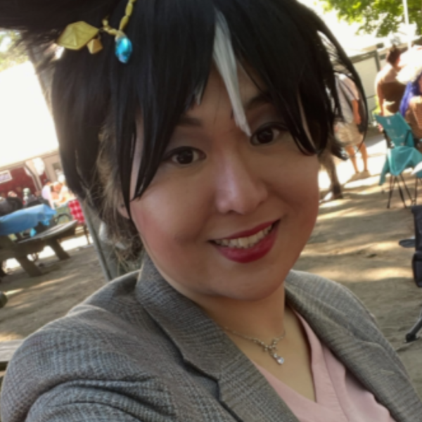 Smiling woman in a gray blazer takes a sunny selfie at an outdoor market or festival.