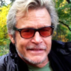 Man with gray hair and stubble wearing dark sunglasses and a black jacket outdoors in a park with autumn trees behind him.