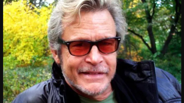 Man with gray hair and stubble wearing dark sunglasses and a black jacket outdoors in a park with autumn trees behind him.