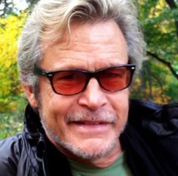 Man with gray hair and stubble wearing dark sunglasses and a black jacket outdoors in a park with autumn trees behind him.