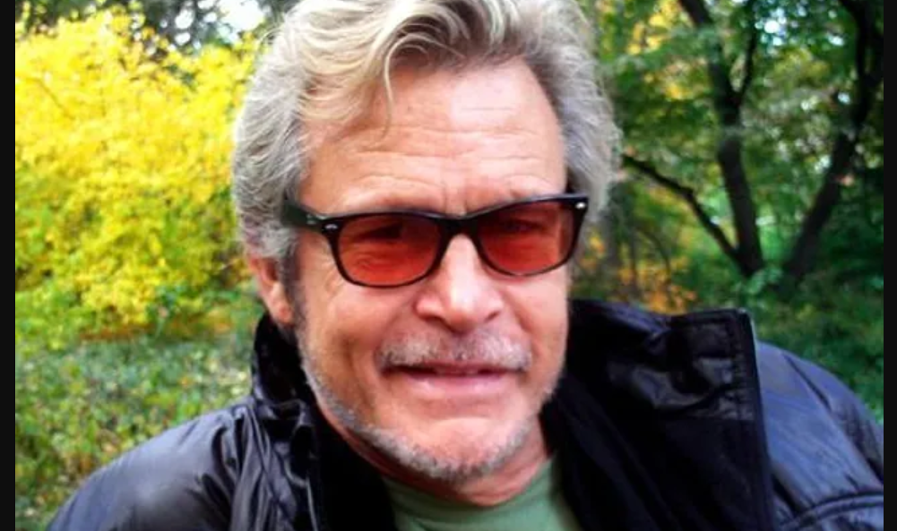 Man with gray hair and stubble wearing dark sunglasses and a black jacket outdoors in a park with autumn trees behind him.